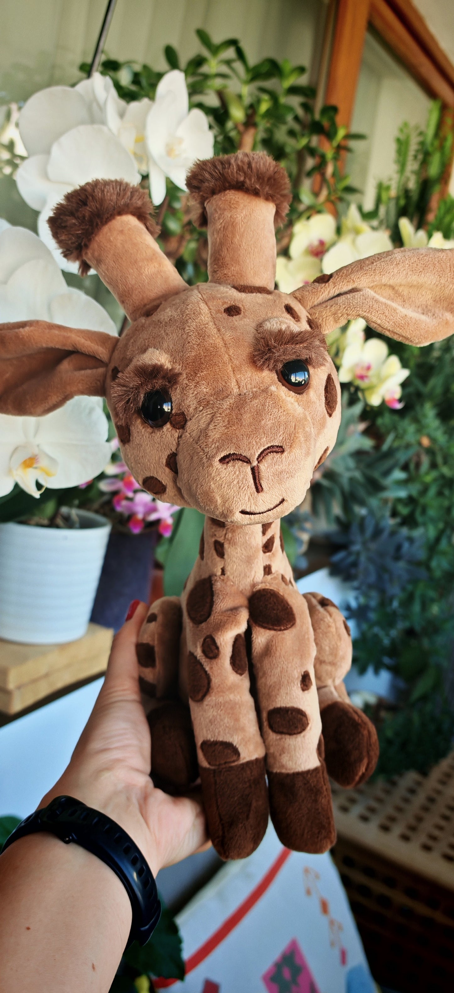Plush giraffe toy held by a hand with a blurred background of plants and flowers.